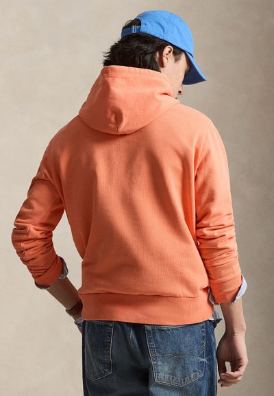 Mann iført en knalloransje hettegenser og blå baseballcap, sett bakfra med hendene nær hoftene, foran en ensfarget beige bakgrunn.