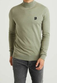 Pull en maille côtelée vert clair avec un col montant et un petit écusson noir sur la poitrine. Coupe ajustée avec manches longues.