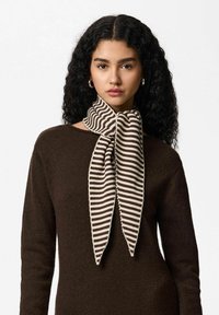 Écharpe tricotée avec un motif à rayures crème et marron, nouée en forme de nœud papillon au niveau du cou. Matière tricotée texturée. Fond de tenue marron.