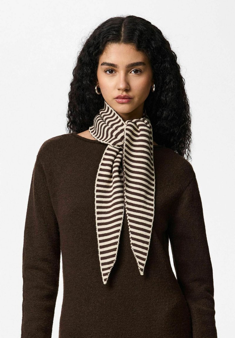 Écharpe tricotée avec un motif à rayures crème et marron, nouée en forme de nœud papillon au niveau du cou. Matière tricotée texturée. Fond de tenue marron.
