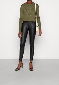 Pullover accorciato verde oliva con design a triangolo texturizzato e borchie, abbinato a pantaloni skinny neri lucidi e tacchi a strappo neri. Accentuato con una borsa piccola.