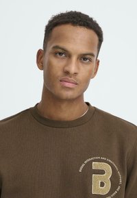 Bruine geribbelde sweatshirt met een ronde hals en een geborduurd cirkel logo met de letter "B" en tekst eronder, contrasterend in lichtbeige.