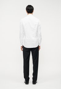 Λευκό πουκάμισο με μακριά μανίκια, ομαλή υφή, κυρτή ούλα και μανσέτες με κουμπιά, σε συνδυασμό με μαύρο παντελόνι, από πίσω.