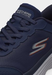 Ναυτικό μπλε αθλητικό παπούτσι Skechers με δίχτυ ύφασμα, υφασμένα κορδόνια, κεντημένο λογότυπο "S" και ετικέτα "Skechers Max Cushion" στη μαξιλαρωμένη γλώσσα.