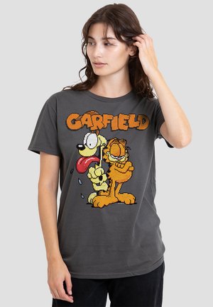 Frau, die sich die Haare richtet und ein graues T-Shirt mit pixelierten Garfield- und Odie-Charakteren sowie dem Text "GARFIELD" darüber trägt.
