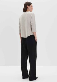 Frau mit glattem braunen Haar, die einen beigen strukturierten Pullover und lockere schwarze Hosen trägt, steht mit den Händen in den Taschen vor einem weißen Hintergrund.