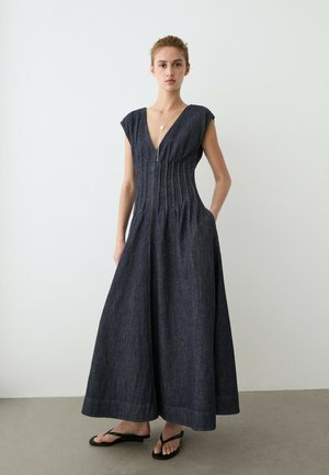 Giovane donna in piedi in una tuta senza maniche blu scuro con gambe larghe e scollo a V profondo, con le mani nelle tasche, indossa sandali neri.
