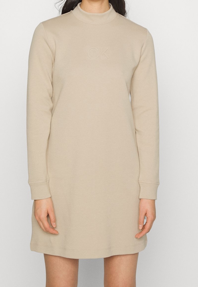 Robe beige à manches longues avec un col haut, présentant un logo "CK" en relief sur la poitrine. Fabriquée en tissu doux et lisse.