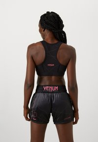 Melna sporta krūšturis ar racerback dizainu, apvienots ar melnām šortiem ar rozā akcentiem un zīmolu. Gluda tekstūra, sportiska piegriezne un minimāla apdare.