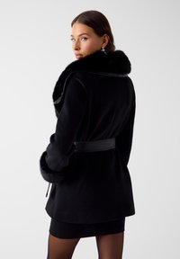 Zwarte jas met een nepbontkraag, een ceintuur in de taille en gestructureerde manchetten. Heeft een gladde stofafwerking en een getailleerd silhouet.