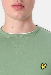 Žalias megztinis su raštuotu apykaklės kraštu ir mažuoju siuvinėjimu geltonos spalvos paukščio logotipu kairėje krūtinės pusėje. Minkšta tekstūra su matomais siuvimo detalėmis.