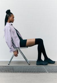 Modèle assis sur une chaise pliante en chrome, portant une chemise oversize rayée de couleur violet clair, un short noir et des chaussettes noires montantes avec des baskets sombres.