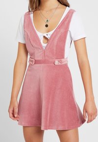 Roze corduroy jurk met een diepe V-hals, nauwsluitend lijfje en uitlopende rok. Aangevuld met twee knopen op de taille voor sluiting.