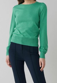 Pull vert en tricot à manches longues avec un col rond et une coupe ajustée. Ourlet et poignets côtelés, porté avec un pantalon bleu marine.