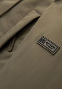 Bežová látka na bundě se hladkým povrchem má tmavý štítek s bílým textem. Detaily zipu a kapes jsou částečně viditelné.