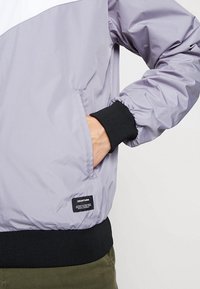 Ljuslila vindjacka med en vit överdel, med elastiska muddarn, en svart ribbad nederkant och en märkt etikett på den nedre vänstra sidan.