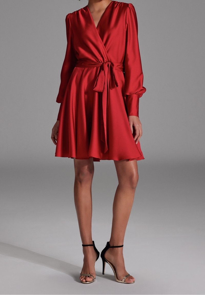 Femme portant une robe portefeuille rouge à manches longues avec une taille nouée et des sandales noires à talons hauts, debout devant un fond gris uni.
