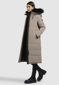 Lys beige puffer-jakke med lang silhuet, pelsforet hætte, lynlås-lukning og sidelommer. Sættes sammen med sorte ankelstøvler.