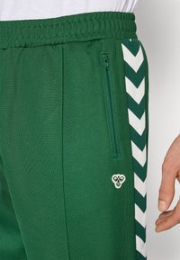 Zöld sportnadrág fehér chevron csíkokkal, cipzáras zsebbel, hímezett logóval és egy látható kézzel a csípő mellett.