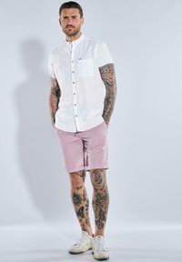 Weißes kurzärmeliges Hemd mit Knöpfen und einer Brusttasche, kombiniert mit rosa Shorts und weißen Turnschuhen. Tattoos an Armen und Beinen sichtbar.