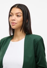 Grønn blazer med en glatt tekstur, åpen frontdesign og strukturerte skuldre, paret med en hvit topp med rund hals under.