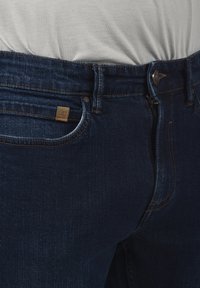 Tamno plave traperice s srednjim strukom, zatvaračem na zip i kopčom na dugme, te malim svijetlosmeđim logotipom na prednjoj džepu.