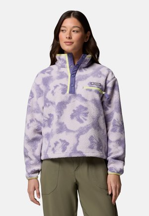 Junge Frau trägt ein lila gepunktetes Fleecepullover mit Druckknöpfen und Reißverschlusstasche auf der Brust, kombiniert mit olivgrünen Hosen, vor weißem Hintergrund.