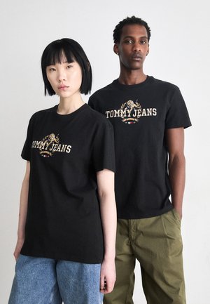 Sorte bomulds T-shirts har "TOMMY JEANS" logo i guld med emblem. Modellen har denim shorts på; baggrunden er neutral.