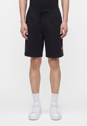 Mann trägt schwarze Shorts mit Kordelzug und goldener Schrift am linken Bein, weiße Socken und weiße Sneakers, steht vor einfachem Hintergrund.