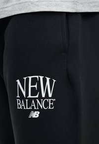 Μαύρο ύφασμα με λευκό κείμενο "NEW BALANCE" και λογότυπο τυπωμένα κοντά στην τσέπη ενός ενδύματος, με γκρι ζώνη μέσης πάνω από.
