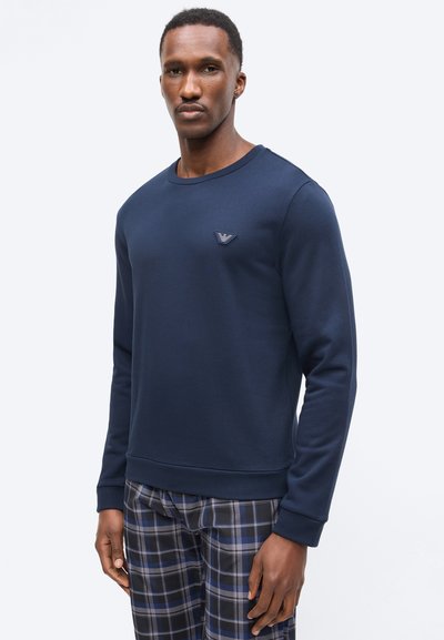 Άνδρας που φοράει μακρυμάνικο φούτερ σε σκούρο μπλε με μικρό λογότυπο στο στήθος και μπλε-γκρι καρό παντελόνι, στέκεται μπροστά σε απλό φόντο.