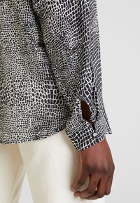 Czarna i biała koszula z wzorem wężowym, z mankietem zapinanym na guziki oraz rozcięciem po boku. Tkanina ma gładką fakturę.