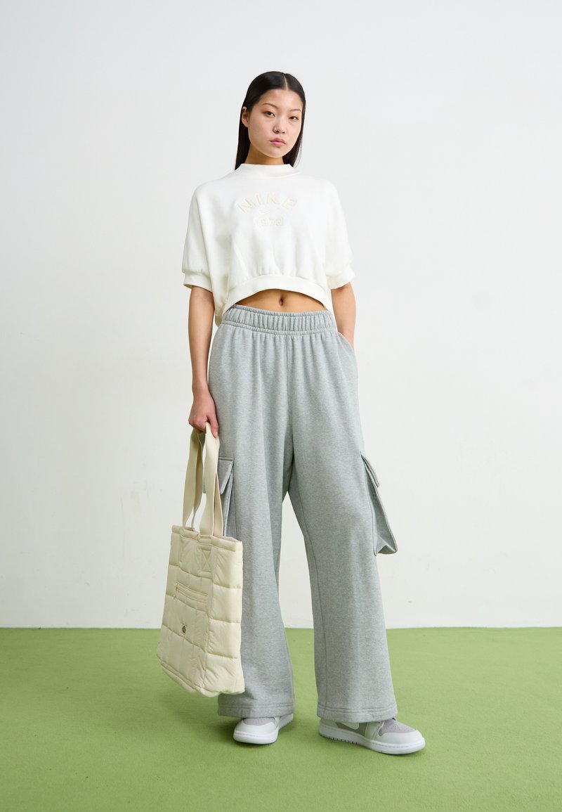 Sweatshirt branco cropped com bordado "NIKE 1972", combinado com calças de fato de treino cinza claro de perna larga com bolsos cargo, e uma mala tote matelassê bege.