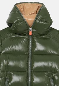 Giacca a vento verde con finitura lucida, grande cappuccio, due tasche laterali con zip e una zip arancione su una fodera interna beige.