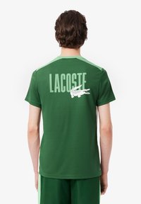 Tricou verde Lacoste cu un design pe umăr de culoare verde deschis, text mare alb "LACOSTE" și un emblema a unui crocodil pe spate.