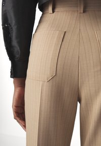 Pantalon beige à fines rayures en tissu léger, avec deux poches arrière et une coupe ajustée, associé à une chemise noire boutonnée.