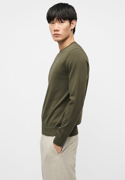 Νέος άνδρας που στέκεται πλαγίως φορώντας ένα πουλόβερ ελιάς και ανοιχτό μπεζ παντελόνι με το ένα χέρι στην τσέπη, μπροστά σε απλό φόντο.