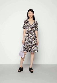 Mulher a usar um vestido de padrão floral com mangas curtas e decote em V, segurando uma mala lilás e calçando sandálias de plataforma pretas.