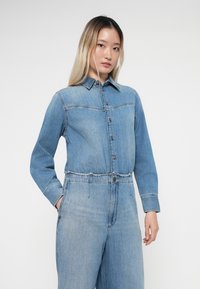 Denim jumpsuit with long sleeves, light blue color, button closure, and an elastic waistband. Features contrast stitching and pockets.