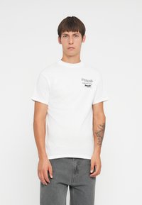 Camiseta de algodón blanca con cuello redondo, mangas cortas y gráficos impresos en negro en el pecho. Combinada con jeans de mezclilla grises. Textura suave.