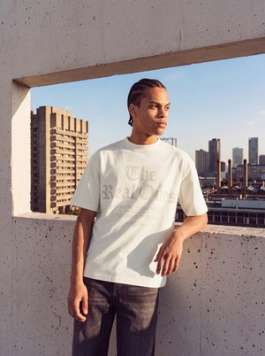 Un homme portant un T-shirt blanc à motif graphique et un jean foncé s’appuie contre un balcon en béton et regarde la silhouette de la ville dans une douce lumière chaude.
