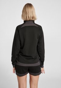 Veste de sport noire avec un col haut, accents gris contrastants et panneaux d'épaules texturés, associée à un short noir.