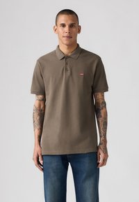 Bruine polo met een kraag en drie knopen, met een klein rood Levi's logo op de linkerkant van de borst. Gemaakt met blauwe jeans.