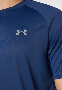 Niebieska sportowa koszulka z krótkim rękawem i szarym logo Under Armour na lewej piersi, noszona przez osobę.