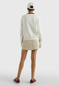 Sudadera beige claro con un cuello negro, combinada con una falda beige. La vista trasera muestra una gorra blanca y unos zapatos con acentos rojos.