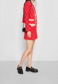 Indumento rosso con motivo a rombi, dettagli crema e bottoni. Abbinato a mocassini neri spessi e a una piccola borsa, in piedi su uno sfondo neutro.