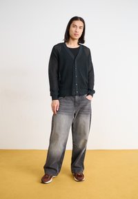 Svart mesh cardigan över en svart skjorta, tillsammans med ljusgrå vida jeans. Bruna och vita sneakers fulländar outfiten. Gul bakgrund.