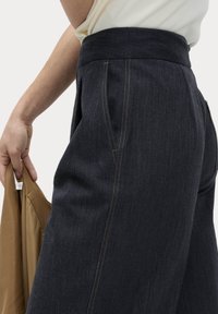 Dunkelblaue Jeanshose mit hohem Bund, ausgestattet mit Seitentaschen und sichtbaren gelben Nähten. In der linken Hand wird ein hellbrauner Stoff gehalten.