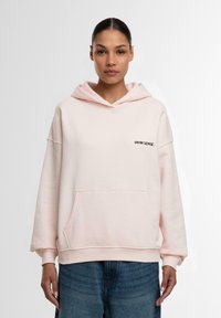 Lys rosa hettegenser laget av mykt stoff, med en frontlomme, ribbede mansjetter og en kontrasterende svart brodert logo på brystet.