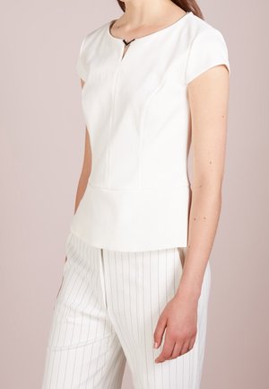 Žena v bílém tričku s krátkým rukávem a malým průstřihem u krku a v bílých kalhotách s tenkými pruhy, stojící před jednobarevným pozadím.